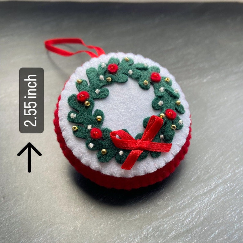 K&ouml;nnte beinhalten: Ein Weihnachtsbaumschmuck aus Filz in Form eines Kranzes mit einer roten Schleife. Der Kranz ist mit gr&uuml;nen Bl&auml;ttern, roten Beeren und goldenen Perlen verziert. Der Schmuck ist 2,55 Zoll im Durchmesser.