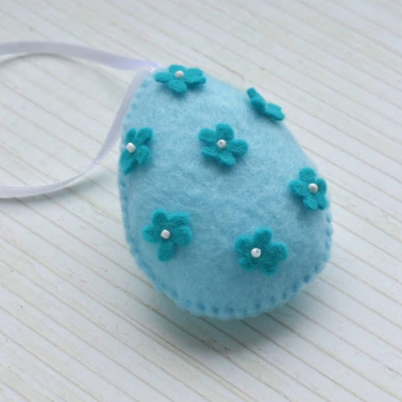 May include: A light blue felt Easter egg ornament with a white ribbon loop and turquoise felt flowers with white centers.
