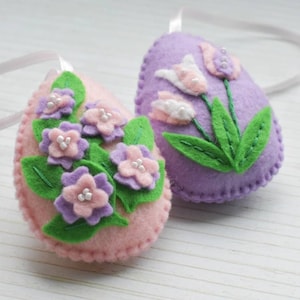May include: Two felt Easter eggs, one pink with purple flowers and green leaves, the other purple with white flowers and green leaves.