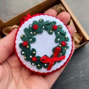 K&ouml;nnte beinhalten: Ein Weihnachtskranz-Anh&auml;nger aus Filz mit rotem Band, gr&uuml;nen Bl&auml;ttern, wei&szlig;en und goldenen Perlen und roten Beeren.