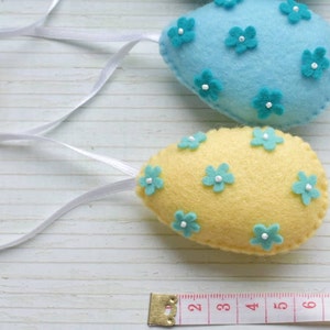 May include: Two felt Easter eggs with blue and yellow fabric and turquoise flower embellishments. The eggs are hanging from white ribbon.