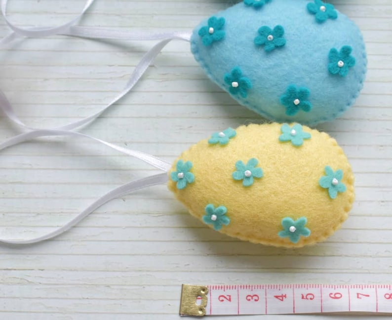 May include: Two felt Easter eggs with blue and yellow fabric and turquoise flower embellishments. The eggs are hanging from white ribbon.