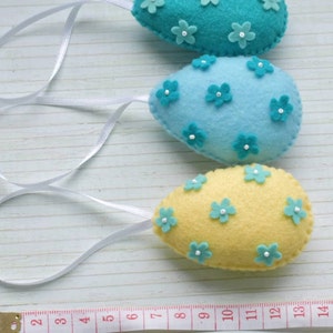 May include: Three felt Easter eggs with blue and yellow colors and white flower embellishments. The eggs are hanging from white ribbon.
