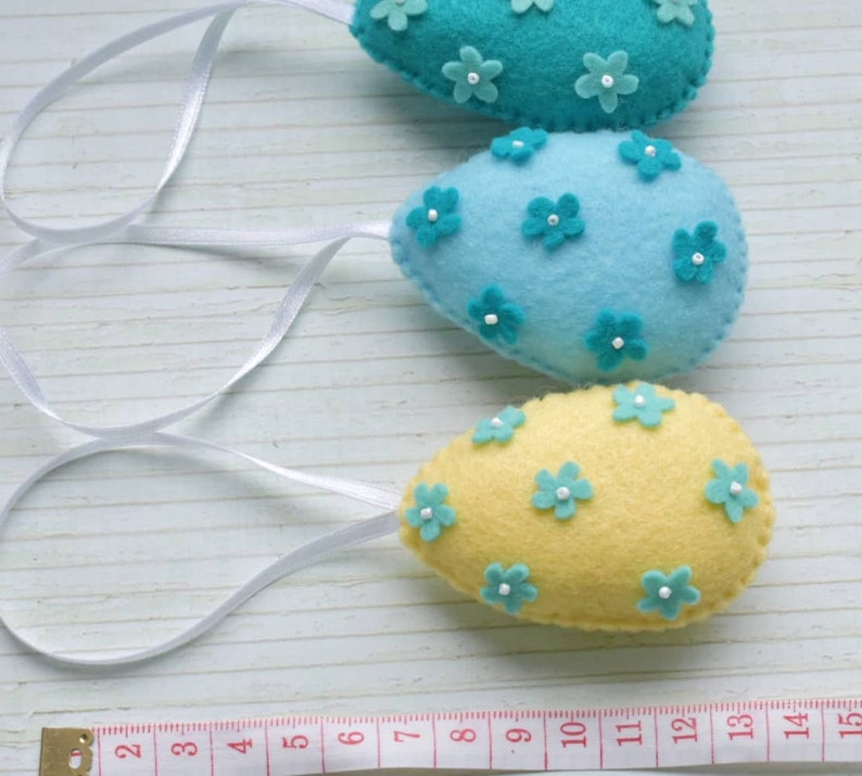 May include: Three felt Easter eggs with blue and yellow colors and white flower embellishments. The eggs are hanging from white ribbon.