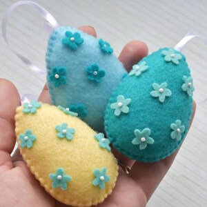 May include: Three felt Easter eggs with white flower embellishments. One egg is light blue, one is teal, and one is yellow.