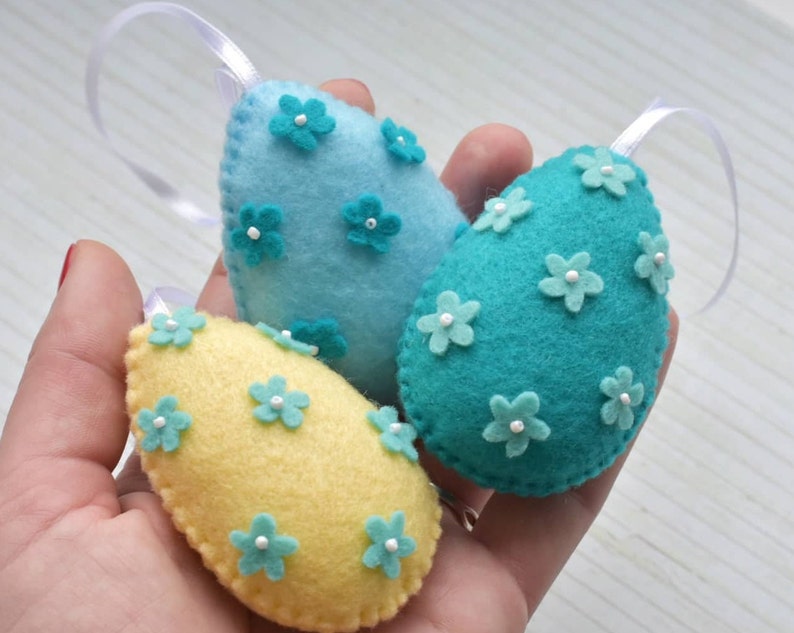 May include: Three felt Easter eggs with white flower embellishments. One egg is light blue, one is teal, and one is yellow.