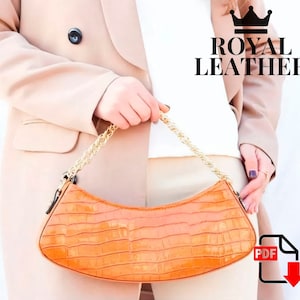 May include: A bright orange handbag with a crocodile-embossed texture and a gold chain strap. The bag is held by a person wearing a beige blazer and white shirt. The text "ROYAL LEATHER" is visible in the image.