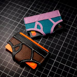 May include: Two leather cardholders in the shape of underwear. One is teal and purple, the other is brown and black. Both have white stitching and hold credit cards. The background is a black grid.