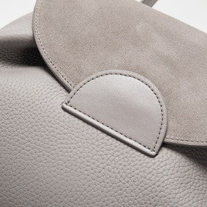 May include: Close-up of a gray leather bag with a suede flap. The flap has a stitched detail in the shape of a half-circle.