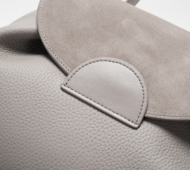 May include: Close-up of a gray leather bag with a suede flap. The flap has a stitched detail in the shape of a half-circle.