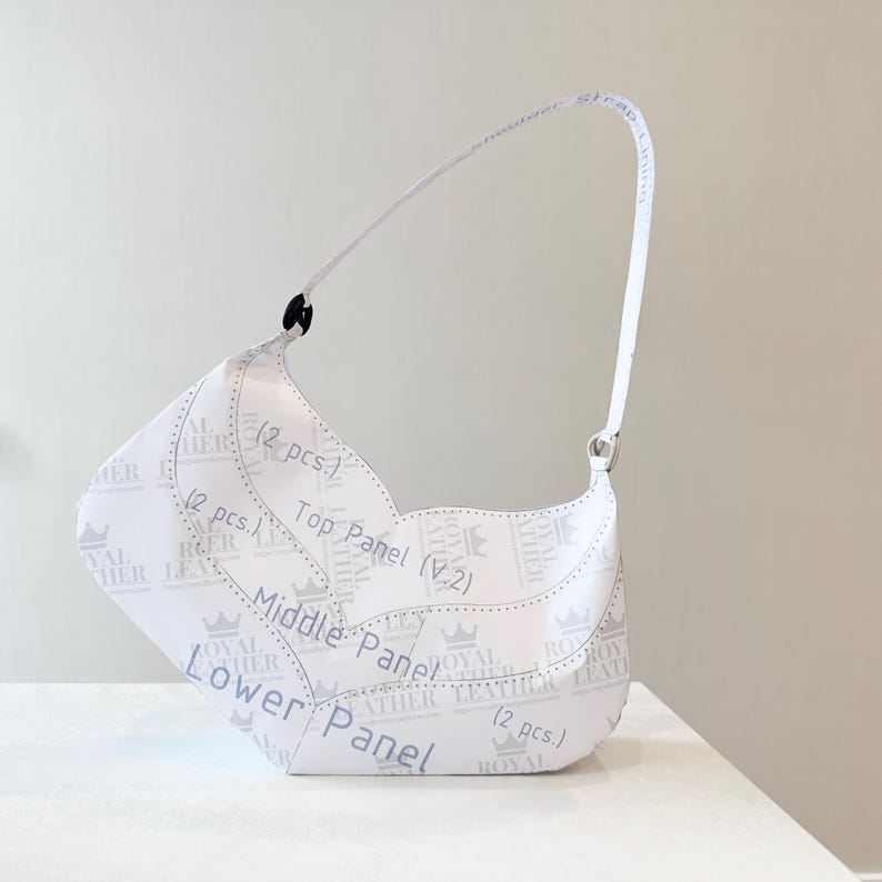 K&ouml;nnte beinhalten: Wei&szlig;e Umh&auml;ngetasche mit geschwungenem Design. Die Tasche hat einen Riemen und ist mit Text und Kronengrafiken bedruckt. Der Text enth&auml;lt "ROYAL LEATHER" und Paneelbeschriftungen. Das Design der Tasche ist sichtbar.