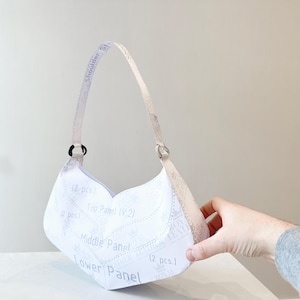 K&ouml;nnte beinhalten: Eine wei&szlig;e Handtasche mit geschwungener Form und Schulterriemen. Der Beutel hat Text, der "Shoulder Strap", "Top Panel", "Middle Panel" und "Lower Panel" lautet. Die Tasche wird von einer Hand gehalten.