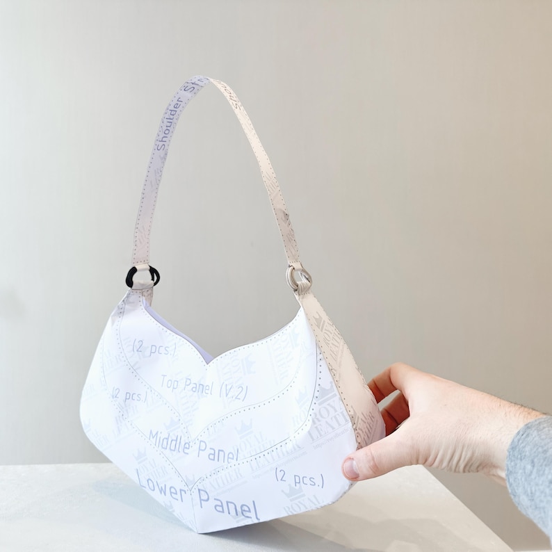 K&ouml;nnte beinhalten: Eine wei&szlig;e Handtasche mit geschwungener Form und Schulterriemen. Der Beutel hat Text, der "Shoulder Strap", "Top Panel", "Middle Panel" und "Lower Panel" lautet. Die Tasche wird von einer Hand gehalten.