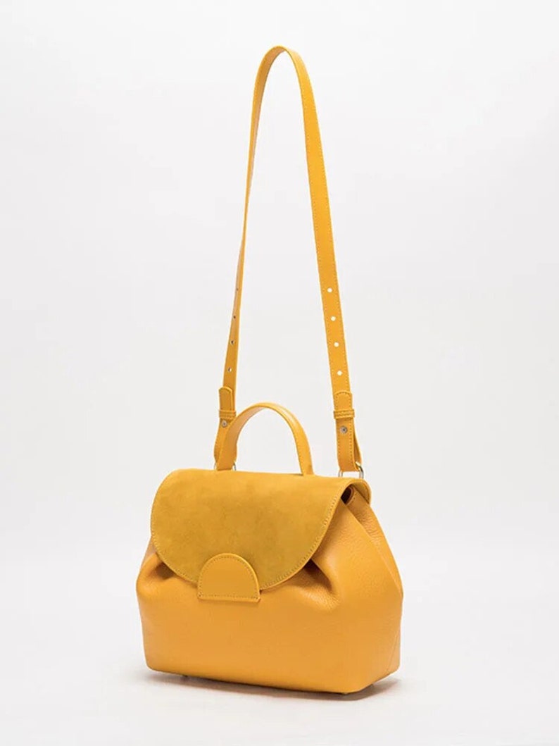 May include: A yellow leather satchel bag with a suede flap and a top handle. The bag has a long adjustable shoulder strap.