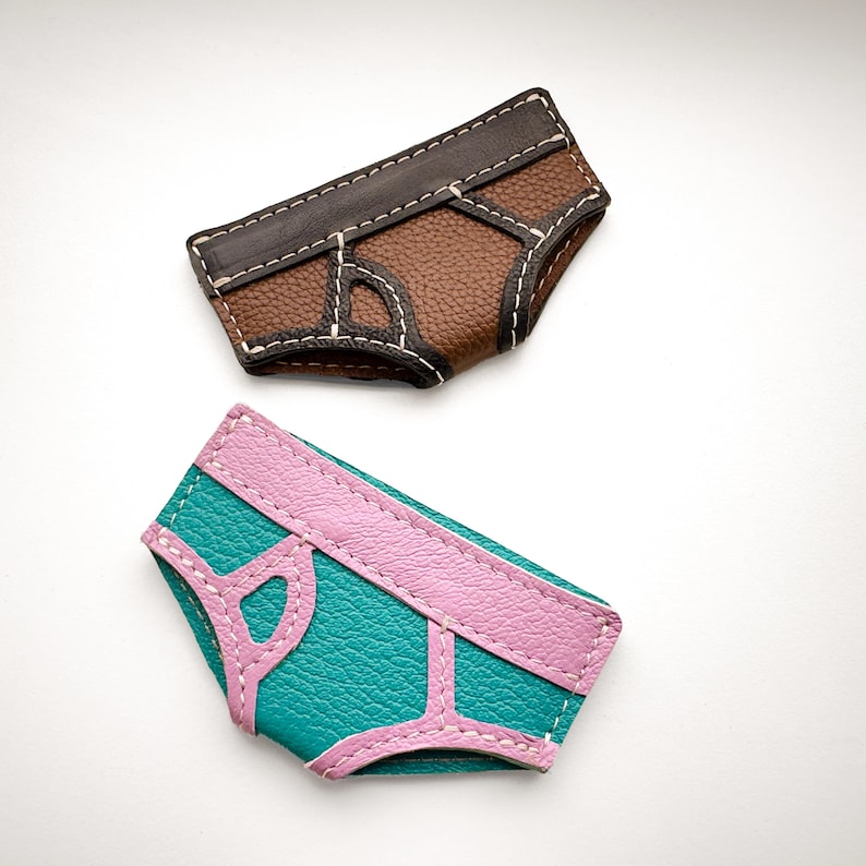 May include: Two leather cardholders shaped like underwear. One is brown with white stitching, the other is teal with pink accents and white stitching. The cardholders are on a white background.