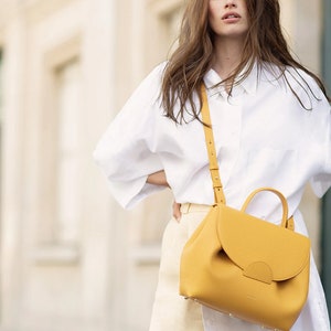 May include: A woman wearing a white shirt and a yellow leather crossbody bag. The bag has a flap closure and a top handle.