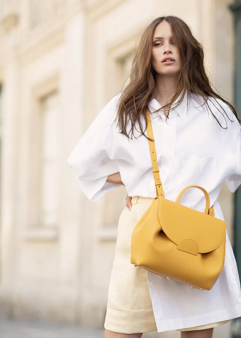 May include: A woman wearing a white shirt and a yellow leather crossbody bag. The bag has a flap closure and a top handle.