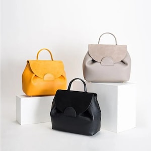 May include: Three leather backpacks in different colors: yellow, gray, and black. Each backpack has a flap with a half-circle cutout and a top handle.