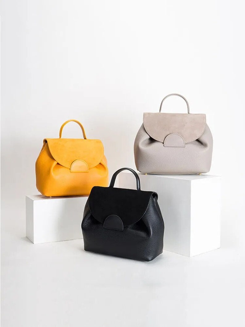 May include: Three leather backpacks in different colors: yellow, gray, and black. Each backpack has a flap with a half-circle cutout and a top handle.