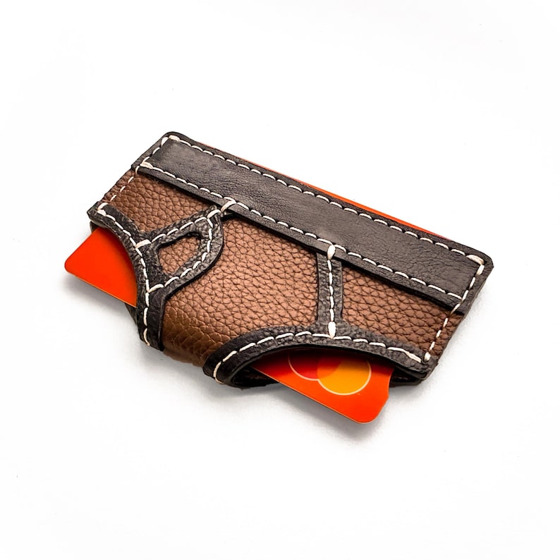 May include: A brown leather card holder with white stitching and a dark brown accent. The card holder has a textured surface and holds a bright orange credit card. The design features a unique stitched pattern.