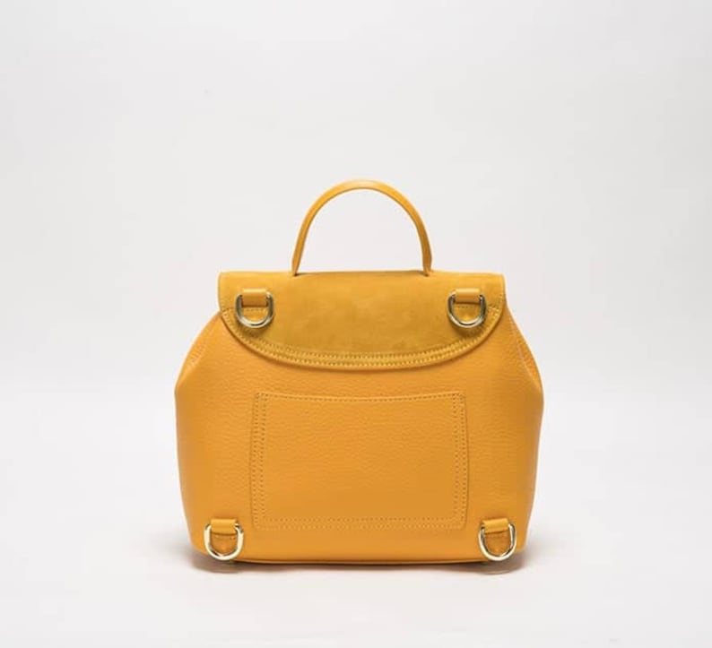 May include: A mustard yellow leather backpack with a suede flap and gold hardware. The backpack has a small pocket on the front and adjustable straps.