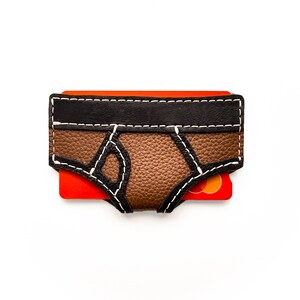 May include: A brown and black leather card holder shaped like a pair of briefs, with white stitching details. An orange credit card is partially visible. The card holder is on a white background.