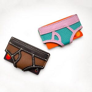 May include: Two leather cardholders in the shape of underwear. One is brown and black with white stitching, the other is teal and pink with orange accents. Each holds a credit card.