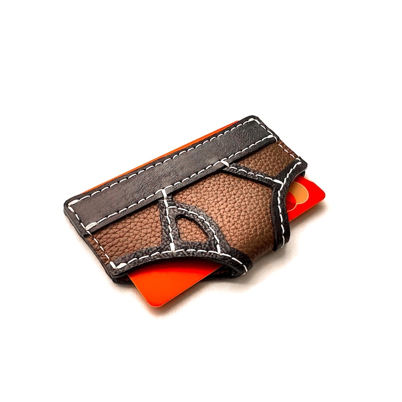 May include: A brown and black leather card holder with white stitching. The card holder has a textured appearance and a rectangular shape. An orange credit card is partially visible.