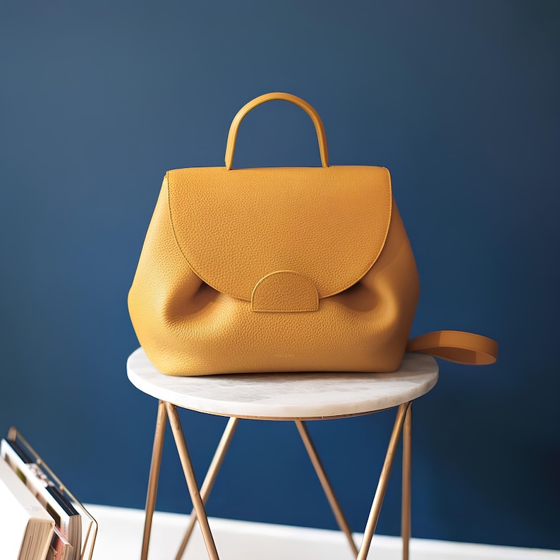 May include: A mustard yellow leather handbag with a curved top and a gold metal handle. The bag is sitting on a white marble table with a gold metal base.