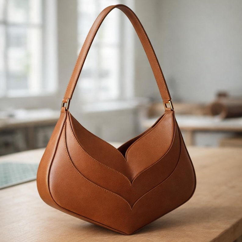 K&ouml;nnte beinhalten: Eine braune Leder-Umh&auml;ngetasche mit einem einzigartigen bl&uuml;tenblattartigen Design. Die Tasche hat einen langen Riemen und goldfarbene Hardware. Die Tasche steht auf einer Holzoberfl&auml;che.