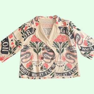 Puede incluir: Chaqueta de manga larga color crema con un estampado único de dragones, flores y texto. El diseño incorpora elementos rojos, negros y verdes. La chaqueta tiene cuello y cierre de botones, ofreciendo una estética vintage.