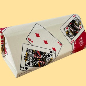 May include: A rectangular, cream-coloured fabric item featuring playing card designs. The cards include the three of diamonds and the queen of spades. The edges are red and white. It is set against a yellow backdrop.