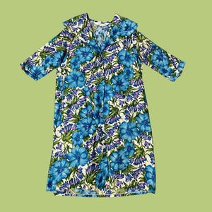 Könnte beinhalten: Ein Vintage-Kleid mit einem auffälligen Blumenmuster. Das Kleid zeigt einen blauen Blumendruck mit weißen und lila Akzenten. Es hat einen Kragen und kurze Ärmel und bietet einen klassischen Retro-Look.