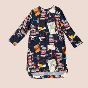 May include: A navy blue dress with a print featuring cameras, film rolls, and other photography-themed elements. The dress has long sleeves and a round neckline. The design is a playful take on a classic dress, suitable for casual wear.