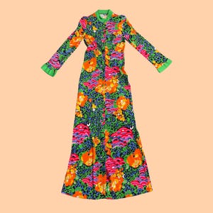 Może przedstawiać: Długa sukienka z długimi rękawami i żywym kwiatowym wzorem. Sukienka ma zielony kołnierz i mankiety, z kolorowym wzorem pomarańczowych, różowych i niebieskich kwiatów na ciemnozielonym tle. Sukienka jest ułożona na płasko.