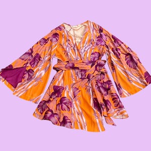Peut inclure: Robe imprimée florale orange et violette de style kimono, avec de larges manches et une ceinture assortie. La robe présente un col en V et une conception portefeuille. Le tissu arbore un motif de feuilles tropicales.
