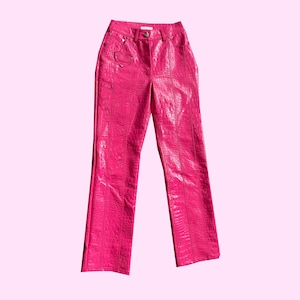 May include: Bright pink, high-waisted trousers with a faux crocodile skin texture. The trousers have a straight leg cut and feature a button closure and a small heart-shaped detail near the pocket. The trousers are made of a shiny material.