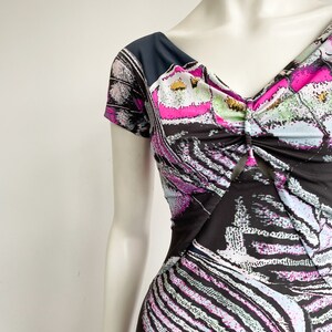 K&ouml;nnte beinhalten: Ein schwarz-wei&szlig; gemustertes Kleid mit rosa und blauen Akzenten. Das Kleid hat einen V-Ausschnitt und kurze &Auml;rmel. Der Stoff ist am Ausschnitt gerafft.
