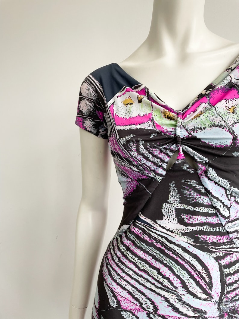 K&ouml;nnte beinhalten: Ein schwarz-wei&szlig; gemustertes Kleid mit rosa und blauen Akzenten. Das Kleid hat einen V-Ausschnitt und kurze &Auml;rmel. Der Stoff ist am Ausschnitt gerafft.