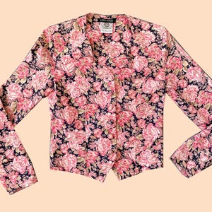 Op de afbeelding: Een blouse met lange mouwen en een bloemenpatroon. De blouse heeft een donkerblauwe achtergrond met grote roze en lichtroze bloemmotieven. De kraag en manchetten zijn zichtbaar en het kledingstuk is dichtgeknoopt.