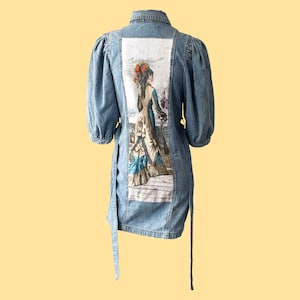 Op de afbeelding: Een lichtblauwe denim shirtjurk met een vintage illustratie van een vrouw in een lange jurk gedrukt op de rug.