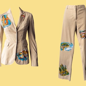 Peut inclure: Ensemble blazer et pantalon beige. Le blazer présente une épingle en forme de ballon de plage et une illustration colorée de deux femmes sur une plage. Le pantalon a plusieurs illustrations de paysages.
