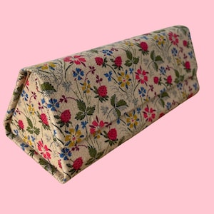 Puede incluir: Un estuche para gafas con estampado floral de forma triangular. El estuche presenta un fondo color crema con un estampado de fresas rojas, flores azules y amarillas y hojas verdes. El estuche está cerrado e inclinado.