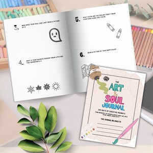 May include: A pink and white journal with the title "The Art & Soul Journal" and the text "365 Days of Creative Prompts". The journal has a colorful paint palette and paintbrush on the cover.