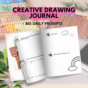 May include: Open journal with black and white illustrations of clouds, a rainbow, and a scroll. The journal has the text "Creative Drawing Journal" and "365 Daily Prompts" on the cover. The journal is open to pages 43, 44, 45, and 46. Page 43 has the text "Combine two of your favorite drawings to create a new one." Page 44 has the text "Take an old drawing and change the." Page 45 has the text "Draw clouds." Page 46 has the text "Draw rainbows in different styles."