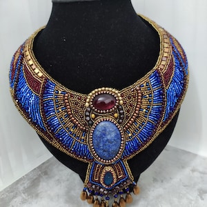 Könnte beinhalten: Eine Halskette mit Bib-Design. Die Halskette besteht aus blauen, goldenen und weinroten Perlen, mit einem großen blauen Stein in der Mitte und einem weinroten Stein oben. Präsentiert auf einer schwarzen Büste.