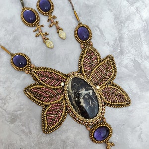 Könnte beinhalten: Ein handgefertigtes Schmuckset mit Halskette und passenden Ohrringen. Die Halskette hat einen großen Mittelstein mit einem schwarz-weißen Muster, umgeben von perlenbesetzten blattartigen Designs in Gold, Rosa und Lila. Die Ohrringe haben ovale lila Steine.