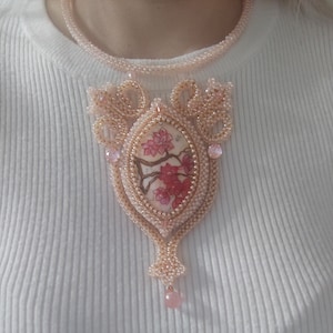 Könnte beinhalten: Eine pfirsichfarbene Perlenkette mit einem Anhänger mit floralem Design. Der Anhänger hat eine ovale Form mit einem gemalten Bild von rosa Kirschblüten auf einem Zweig. Die Halskette umfasst ein passendes Perlen-Choker und eine hängende rosa Perle.