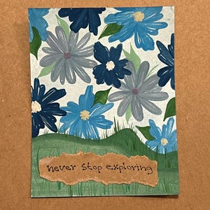 Può includere: Piccolo dipinto rettangolare con fiori blu e grigi con foglie verdi su sfondo bianco. In basso, un'area erbosa verde con la scritta "never stop exploring" su una striscia di carta marrone.