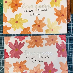 May include: Two rectangular cards featuring hand-painted floral patterns. The top card has yellow and orange flowers, while the bottom card has pink and orange flowers. Both cards display the text "love yours snail mail club" in the center.
